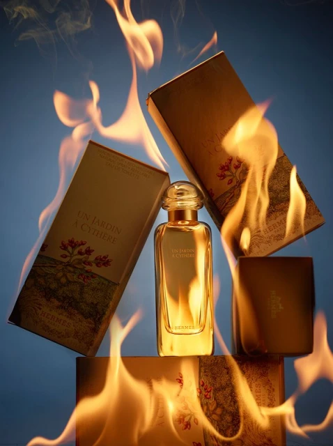 Gold Hermès 'Un Jardin à Cythère' perfume bottle surrounded by matching packaging and flames against a gradient blue background.