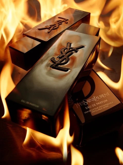 Three gold YSL perfume bottles are arranged in front of flames, creating a dramatic and eye-catching beauty product shot.