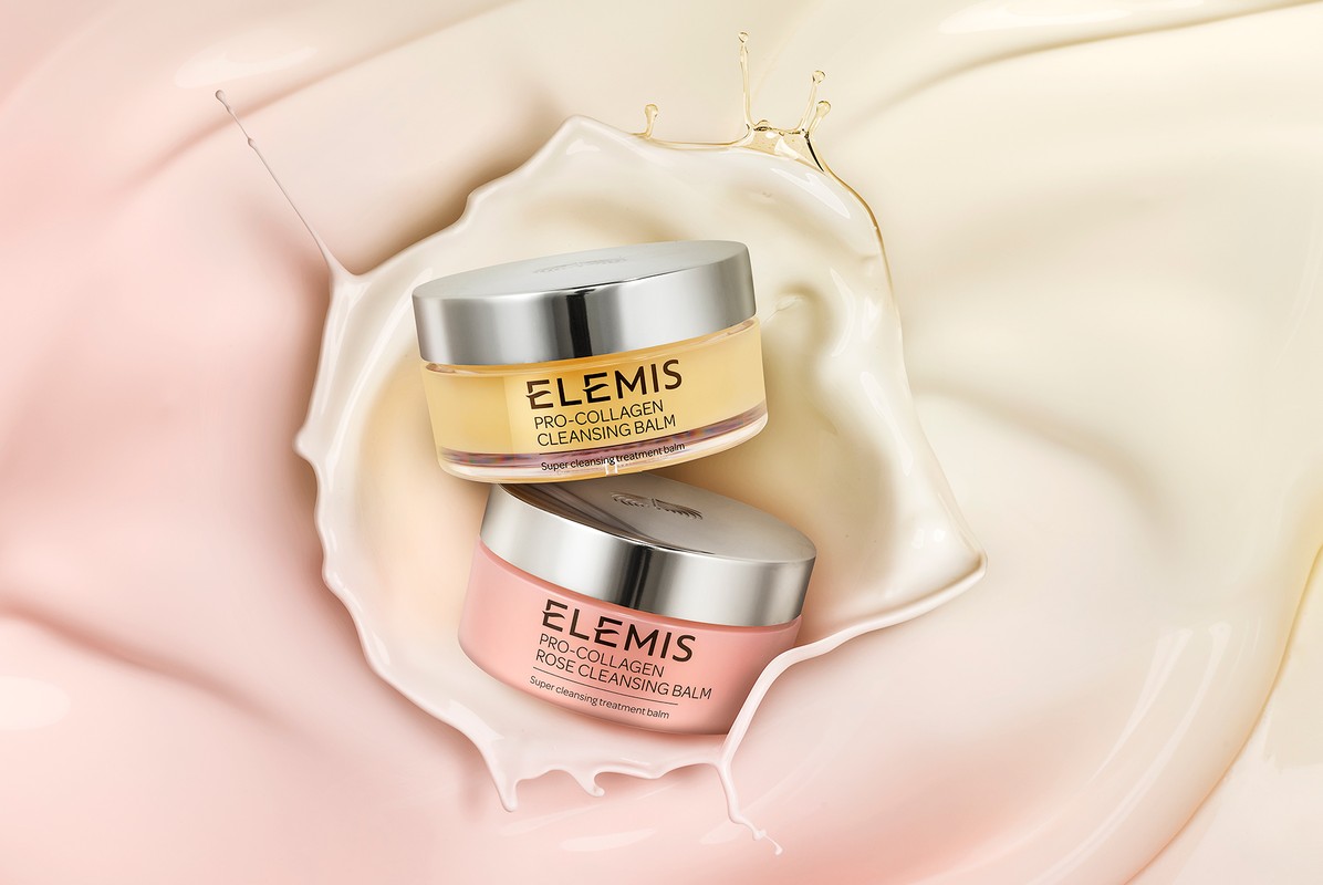 Two jars of Elemis skincare products sit stacked against a cream and light pink background with a splash of creamy liquid.