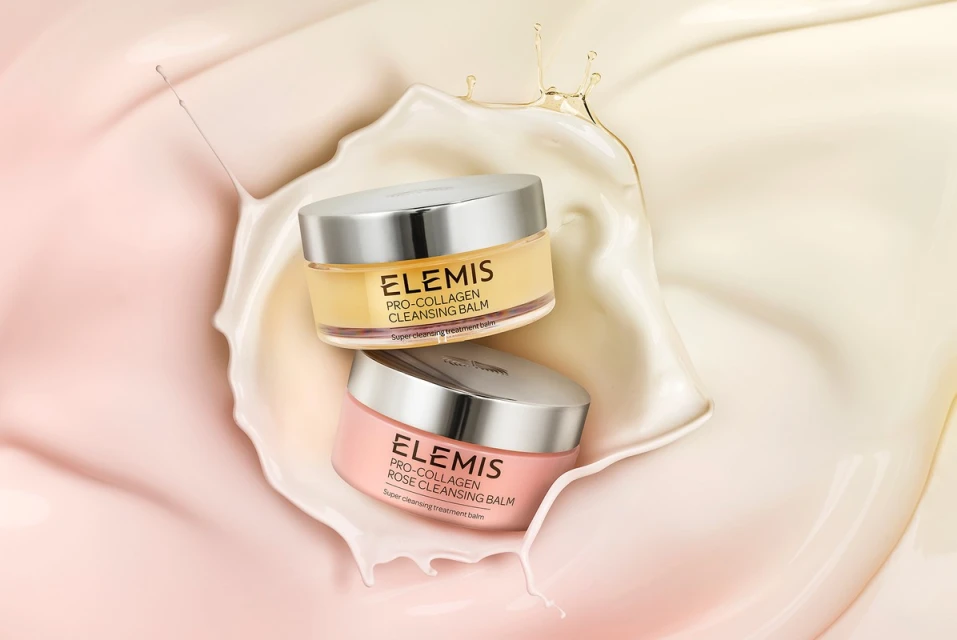 Two jars of Elemis skincare products sit stacked against a cream and light pink background with a splash of creamy liquid.