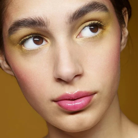 Close-up portrait of a woman with yellow eyeshadow and glossy pink lips against an ochre background.