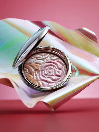 Silver compact containing pink and gold powder embossed with a rose pattern on a colorful iridescent surface against a pink backdrop.