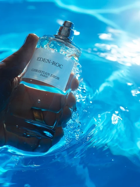 A hand holds a bottle of Eden-Roc Christian Dior perfume underwater against a bright blue background with water droplets and ripples.