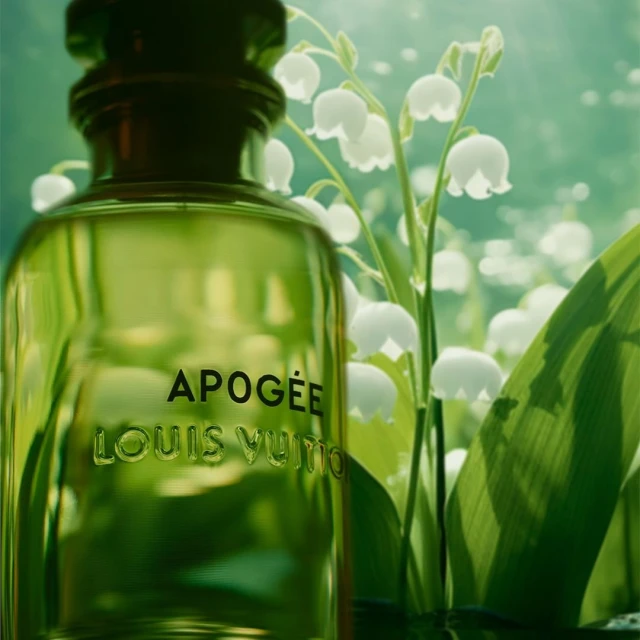 Close-up of a green glass Apogée Louis Vuitton perfume bottle with white lily of the valley flowers and green leaves behind it.
