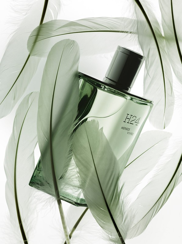 Close-up of a green glass perfume bottle with a black cap, surrounded by white feathers on a white background. Studio lighting.