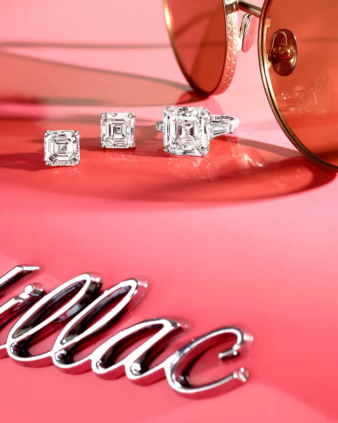 Close-up studio shot of diamond earrings and ring beside sunglasses and partial logo on pink background, jewelry product photography.