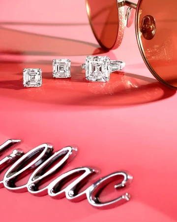Close-up studio shot of diamond earrings and ring beside sunglasses and partial logo on pink background, jewelry product photography.