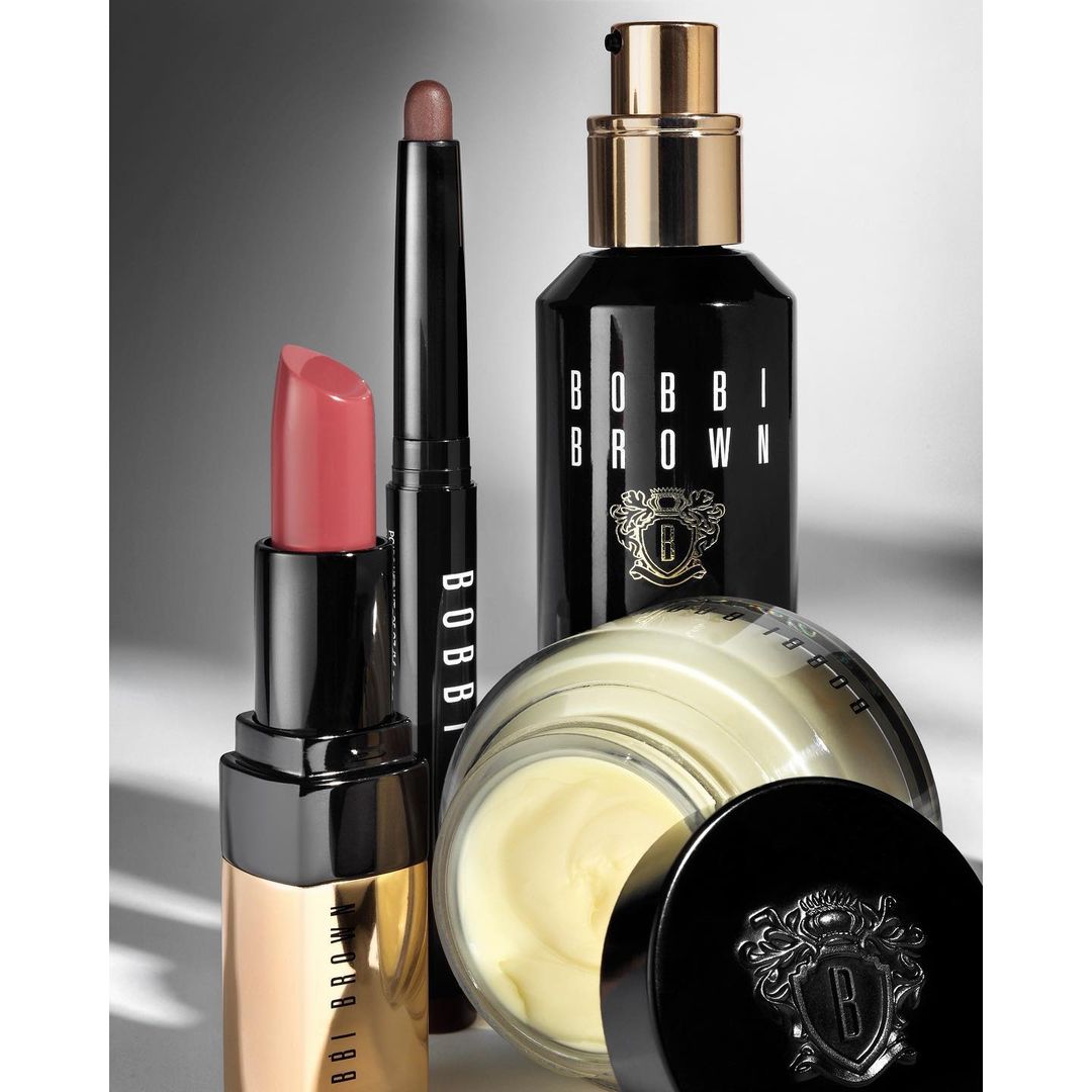 Bobbi Brown beauty products: lipstick, eyeshadow stick, facial serum, and face cream, all in sleek black and gold packaging.