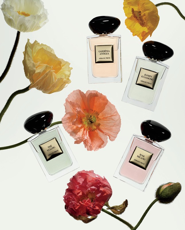 Five perfume bottles are arranged among pink, orange, yellow and white poppies with green stems against a bright white background.