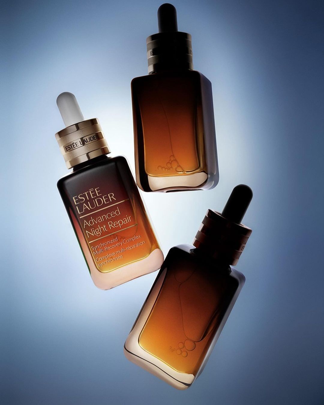 Three Estee Lauder Advanced Night Repair serum bottles arranged at angles against a graduated blue background with soft lighting.