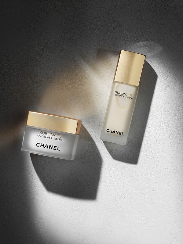 Chanel Sublimage skincare products, La Crème Lumière and L'Essence Lumière, sit on a textured white surface in a beauty still life.