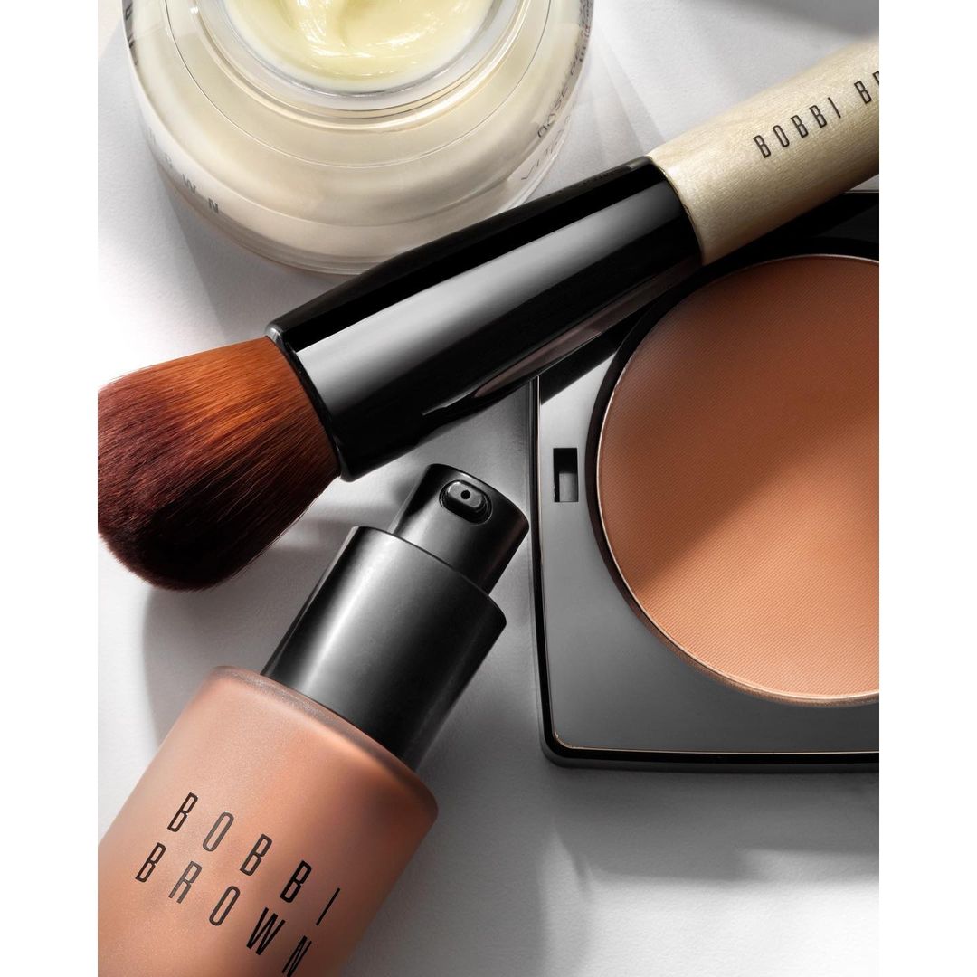 Close-up studio shot of Bobbi Brown makeup products including foundation, powder, and brush on white background for beauty editorial.