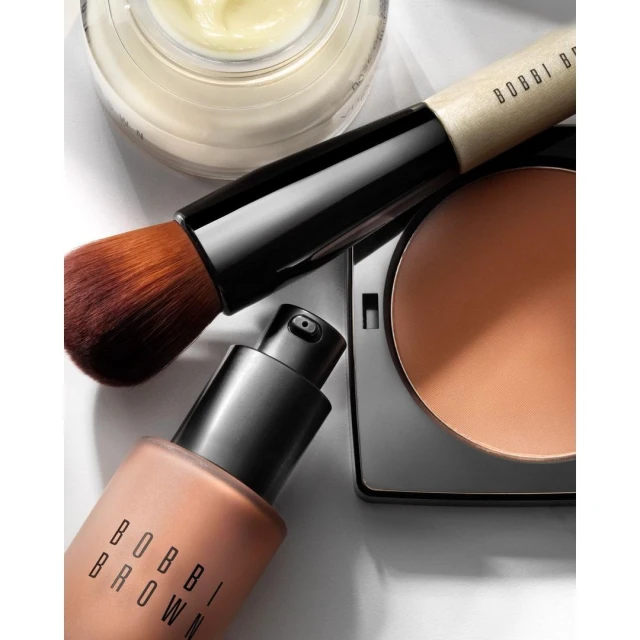 Close-up studio shot of Bobbi Brown makeup products including foundation, powder, and brush on white background for beauty editorial.