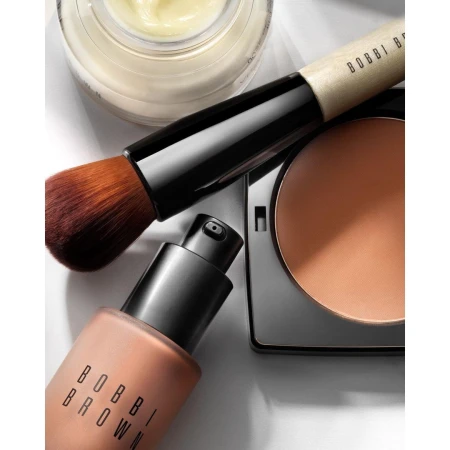 Close-up studio shot of Bobbi Brown makeup products including foundation, powder, and brush on white background for beauty editorial.