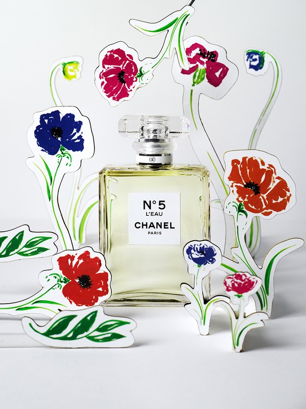 Chanel No. 5 L'EAU perfume bottle stands on a white surface surrounded by colorful painted paper flower cutouts.