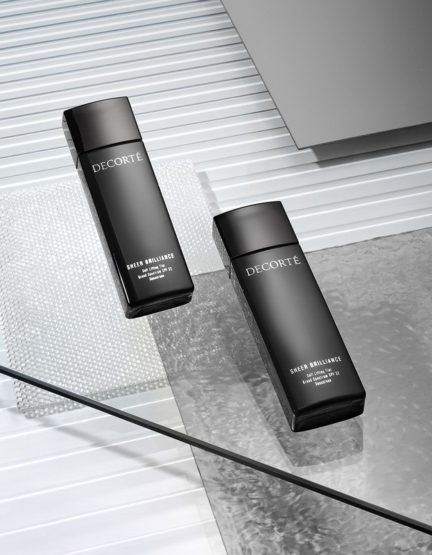 Two black bottles of Decorté Sheer Brilliance sit on a reflective surface against a horizontally lined gray background in a beauty product shot.