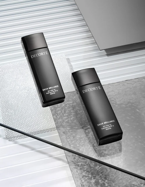 Two black bottles of Decorté Sheer Brilliance sit on a reflective surface against a horizontally lined gray background in a beauty product shot.