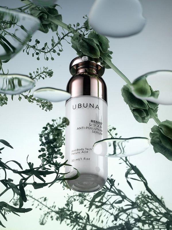 Cosmetic serum bottle by Ubuna in studio product shot with botanical sprigs and water droplets, white bottle with gold accents.