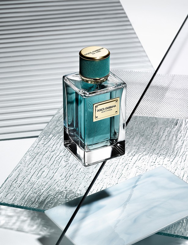 Turquoise Dolce & Gabbana Velvet Treasure eau de parfum in a clear glass bottle with a gold cap, on reflective, geometric surfaces.
