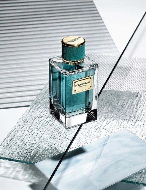 Turquoise Dolce & Gabbana Velvet Treasure eau de parfum in a clear glass bottle with a gold cap, on reflective, geometric surfaces.