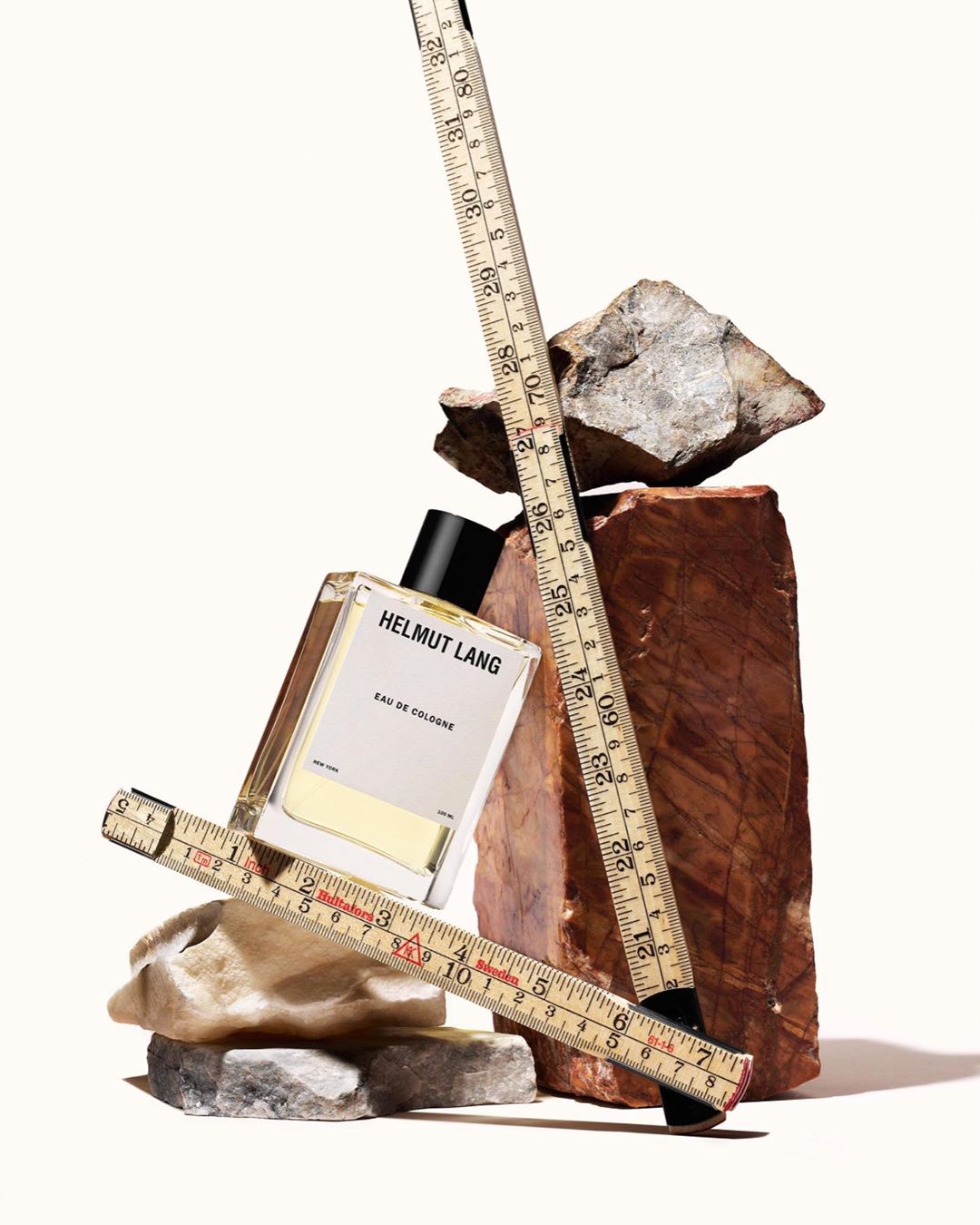 Cologne bottle by Helmut Lang sits amid rocks and measuring sticks. Modern fragrance still life in minimalist aesthetic, studio shot.