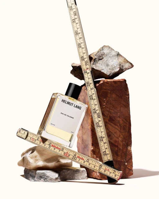 Cologne bottle by Helmut Lang sits amid rocks and measuring sticks. Modern fragrance still life in minimalist aesthetic, studio shot.