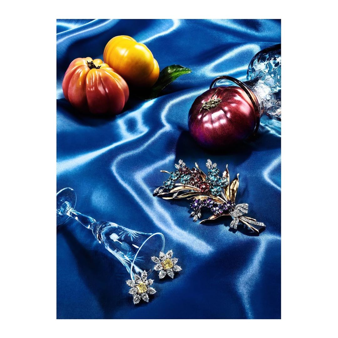 Still life photo of jewelry, tomatoes, and a glass on a blue silk background. A pair of earrings are in a glass, and a brooch lies nearby.