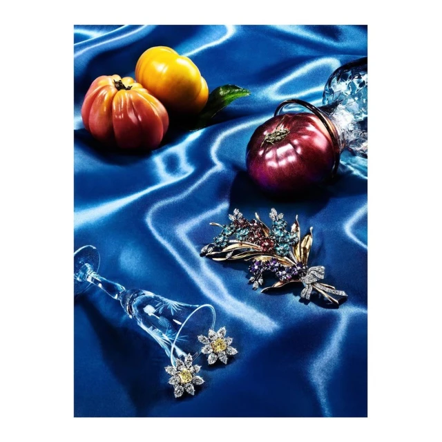 Still life photo of jewelry, tomatoes, and a glass on a blue silk background. A pair of earrings are in a glass, and a brooch lies nearby.