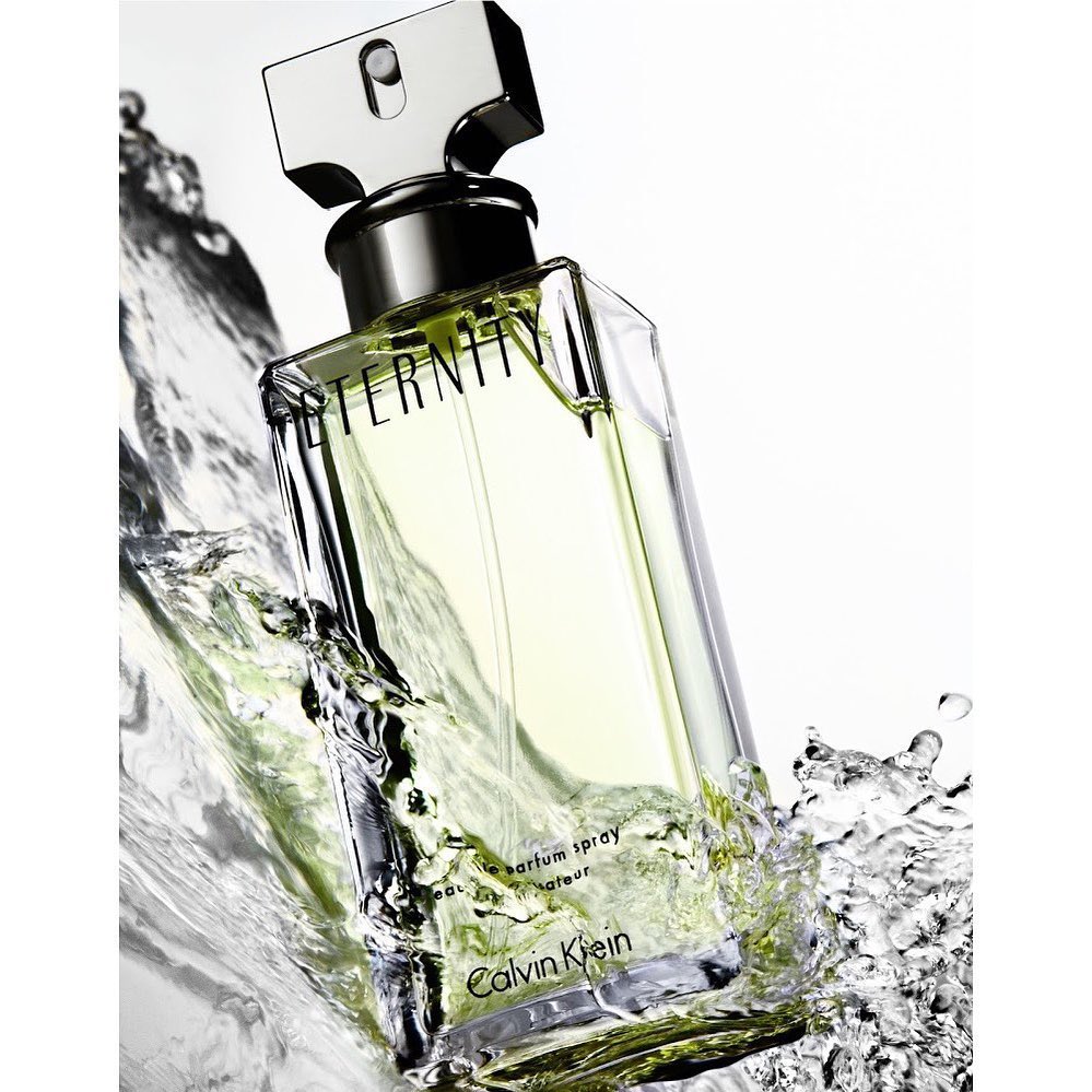 Close-up of a Calvin Klein Eternity perfume bottle with silver accents, surrounded by splashing water against a white background.