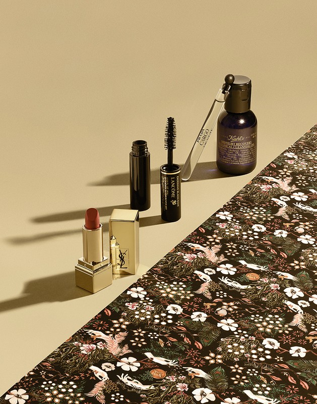 Still life of lipstick, mascara, essence, and skincare on floral patterned paper and beige surface in a product photography composition.