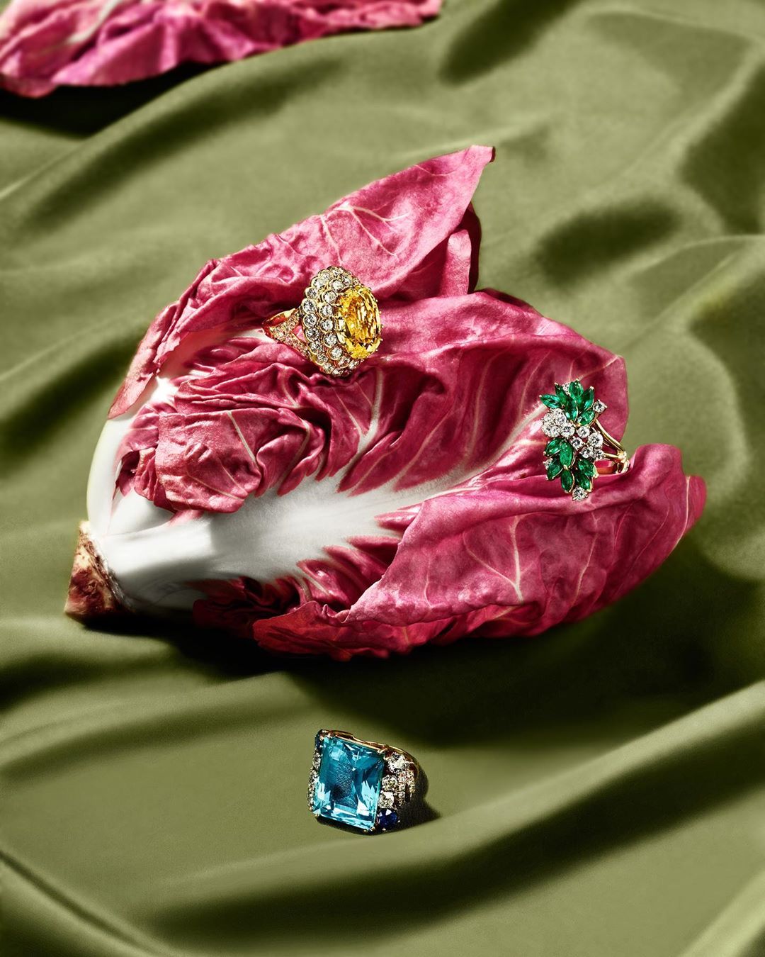 Three jeweled rings are arranged on a leaf against an olive green fabric backdrop in a bright, well-lit, eye-level still life photograph.