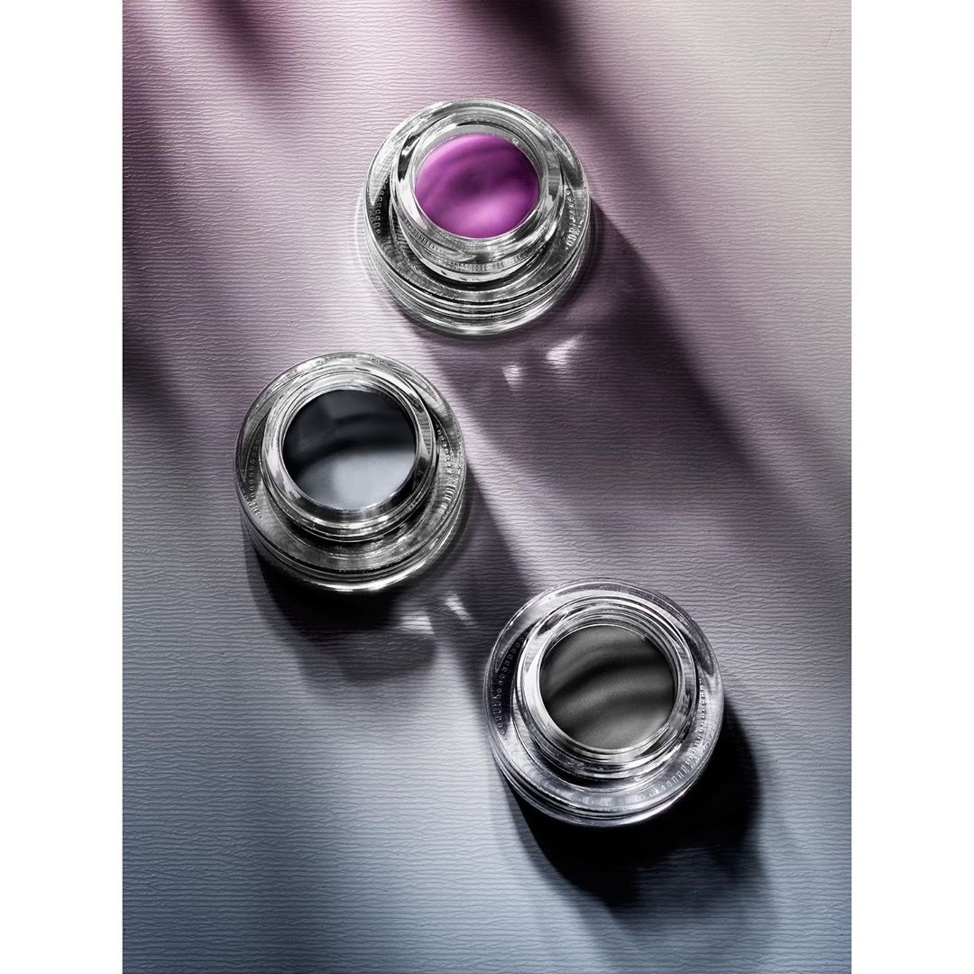 Three clear glass cosmetic containers sit on a purple and grey textured surface under direct lighting, creating sharp shadows.