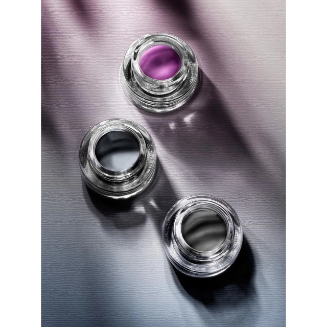 Three clear glass cosmetic containers sit on a purple and grey textured surface under direct lighting, creating sharp shadows.