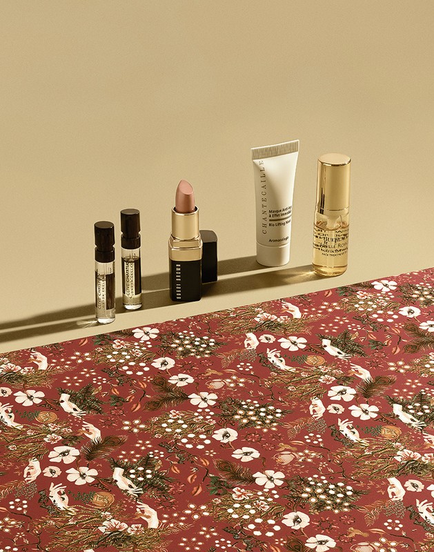 Cosmetics product still life: lipstick, lotions, and perfume bottles displayed on a red floral fabric against a tan background.