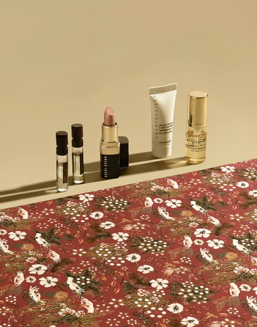 Cosmetics product still life: lipstick, lotions, and perfume bottles displayed on a red floral fabric against a tan background.