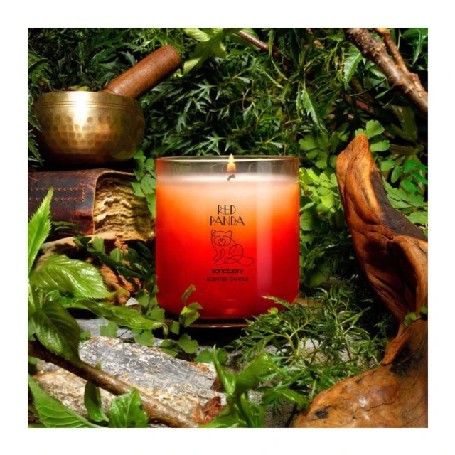 Aromatic red candle with a visible flame surrounded by verdant foliage, a brass bowl, and a vintage book in an indoor setting.