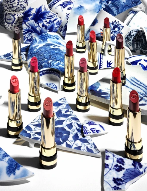 Close-up of several tubes of lipstick, arranged artfully on a white surface amid shattered pieces of blue and white porcelain.