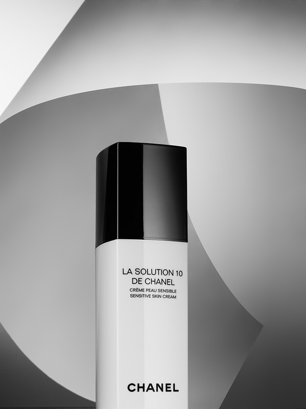 Product shot of a Chanel 'La Solution 10 de Chanel' cream bottle in black and white, set against a backdrop of curved paper.