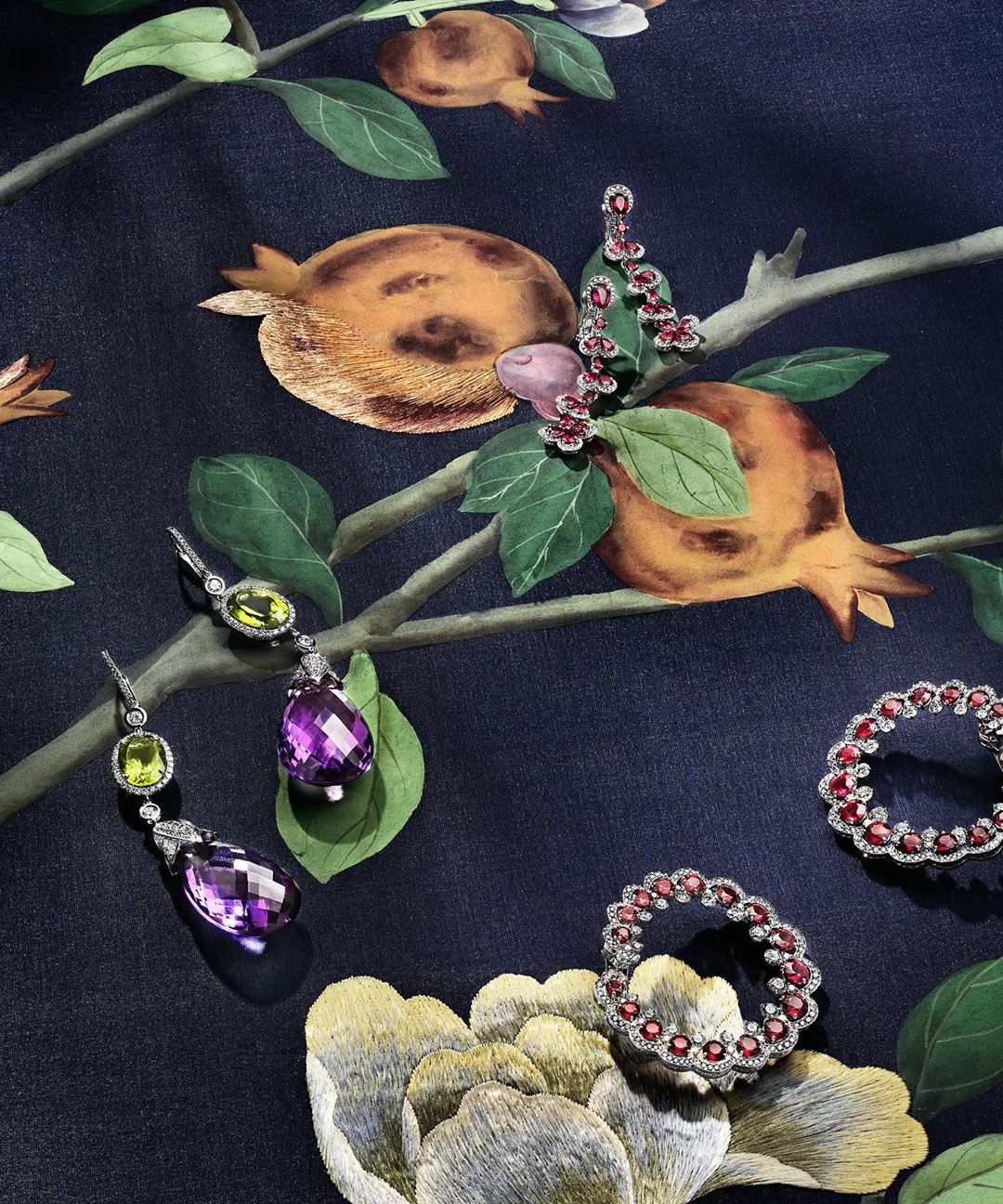 A floral-patterned fabric backdrop displays high-end earrings and rings with colorful gemstones, in a flat lay studio product shot.