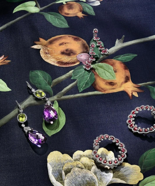 A floral-patterned fabric backdrop displays high-end earrings and rings with colorful gemstones, in a flat lay studio product shot.