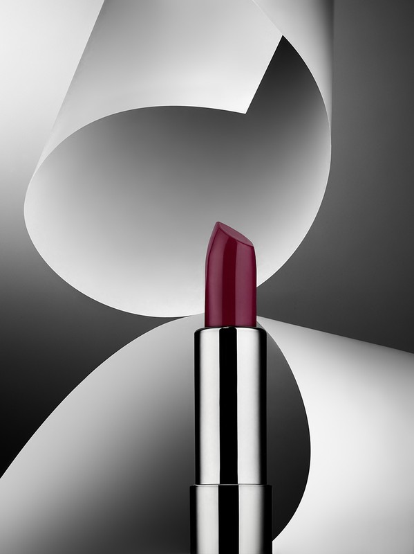 Close-up studio shot of a silver lipstick tube with a dark red shade against a backdrop of gray and white abstract paper shapes.