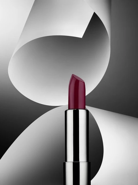 Close-up studio shot of a silver lipstick tube with a dark red shade against a backdrop of gray and white abstract paper shapes.