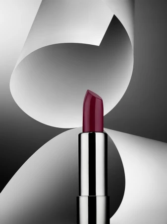Close-up studio shot of a silver lipstick tube with a dark red shade against a backdrop of gray and white abstract paper shapes.