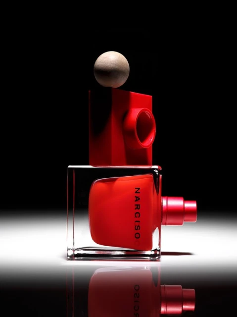 Product shot of Narciso Rodriguez fragrance: a red bottle with a clear cube base, sitting on a reflective surface with a black backdrop.