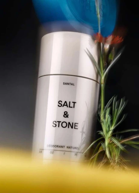 A white cylindrical deodorant container by Salt & Stone is featured next to a green plant with a black and gold background.