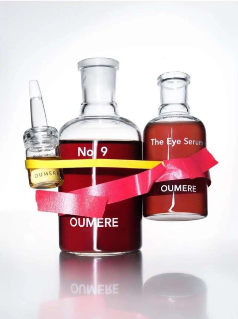 Three skincare bottles of different shapes with red and yellow ribbons on a white background. OUMERE brand visible on all bottles.