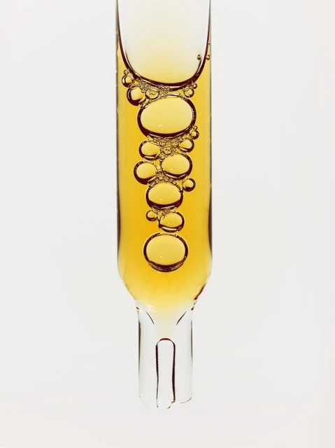 Close-up of a transparent glass pipette filled with golden liquid with bubbles on a plain white background, pharmaceutical or beauty product.
