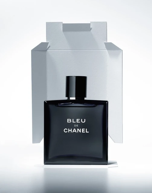 Square bottle of Bleu de Chanel perfume with a black cap in front of a white box against a white background. Studio lighting.