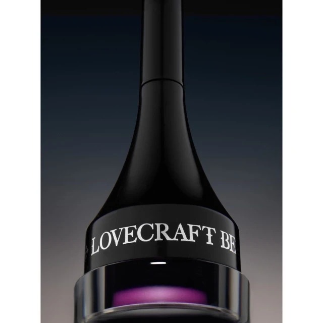 Close-up studio shot of a black cosmetic container with purple liquid at the base. The brand name 'LOVECRAFT BE' is displayed.