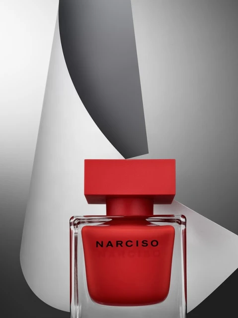 Close-up of a red Narciso Rodriguez perfume bottle with a square cap, set against a grey and white paper background.